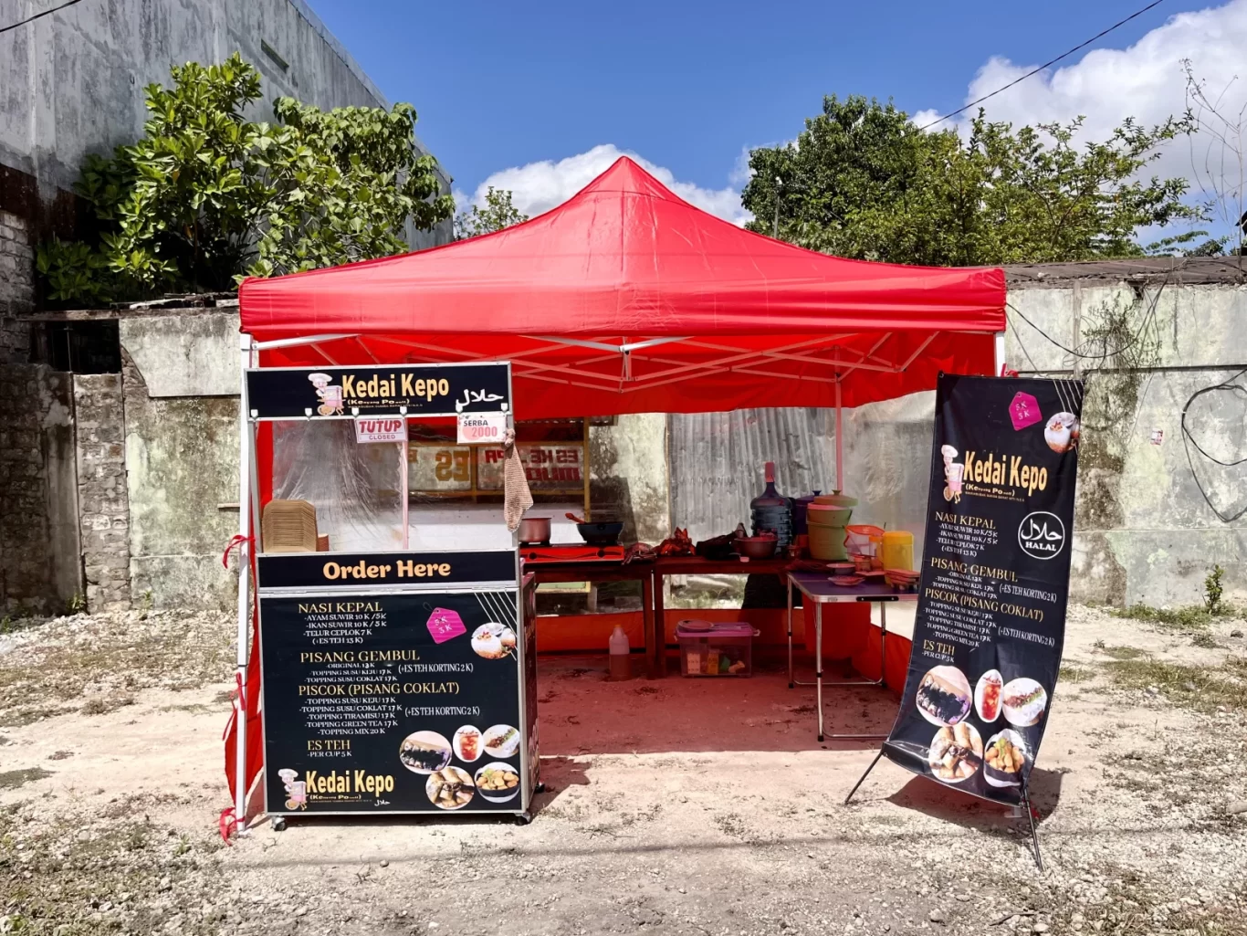 A red tent food stall named "Kedai Kepo" with black and white menus stands on a dirt patch against a concrete wall and green trees under a blue sky.