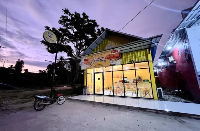 A brightly lit yellow Xie-Xie cafe building with large glass windows stands at dusk under a purple sky, with a motorcycle parked outside and a dark tree behind it.