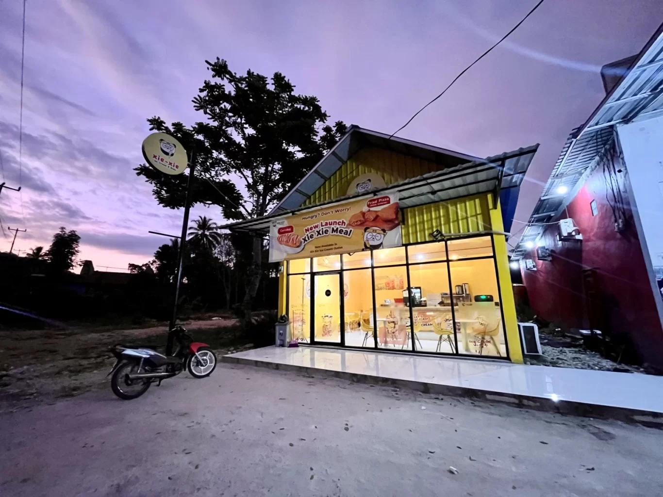 A brightly lit yellow Xie-Xie cafe building with large glass windows stands at dusk under a purple sky, with a motorcycle parked outside and a dark tree behind it.