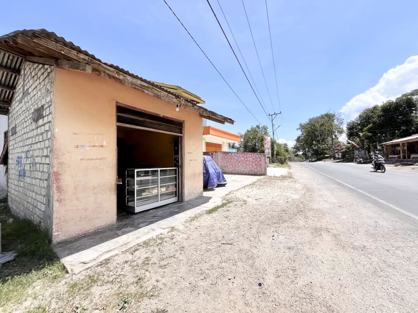 A small, peach-colored food stall with a glass display case stands beside a dirt road and paved street under a bright blue sky in Sumba.