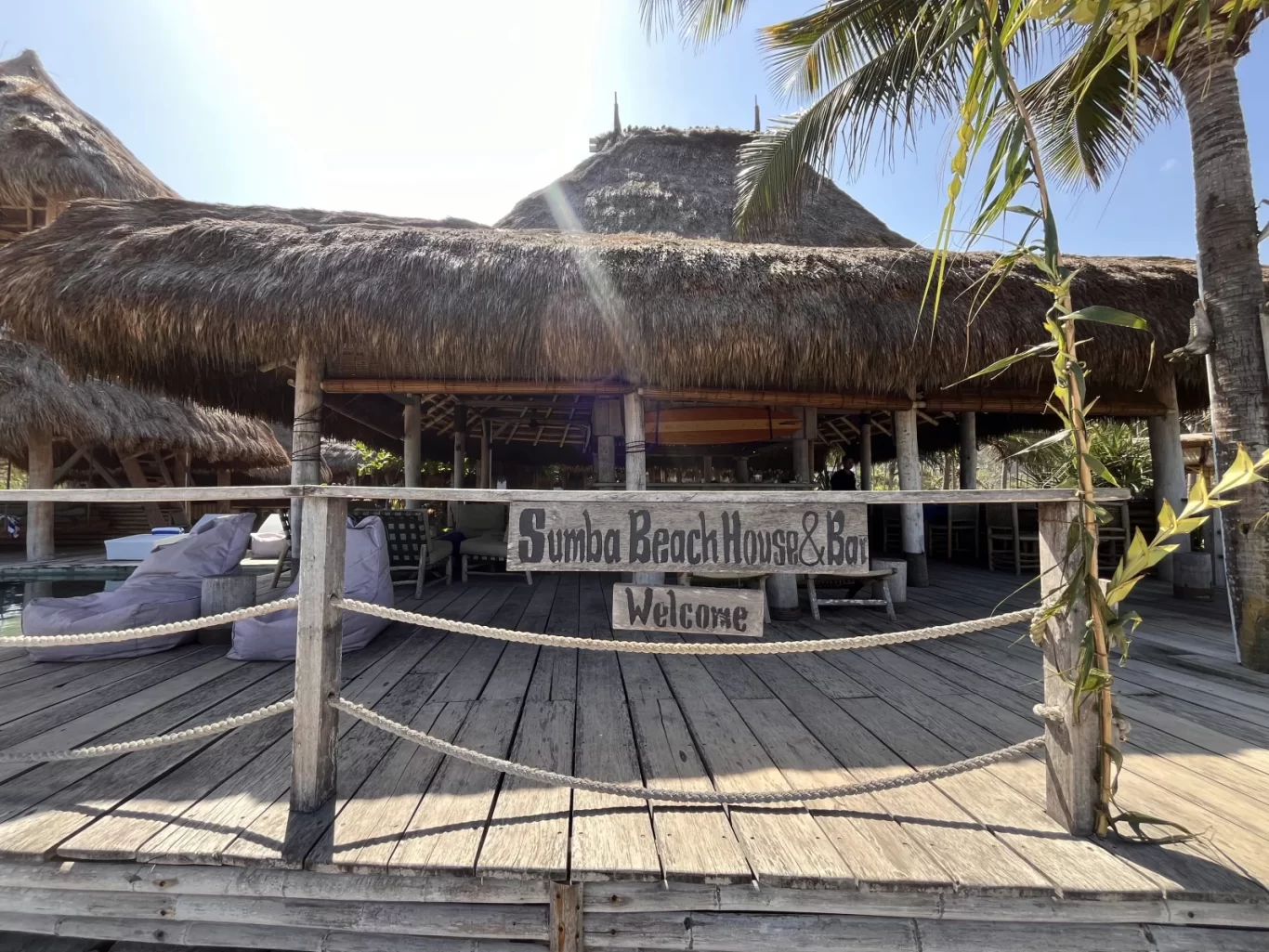 A rustic Sumba Beach House & Bar building with a thatched roof stands on a wooden deck under a bright blue sky, with a "Welcome" sign.