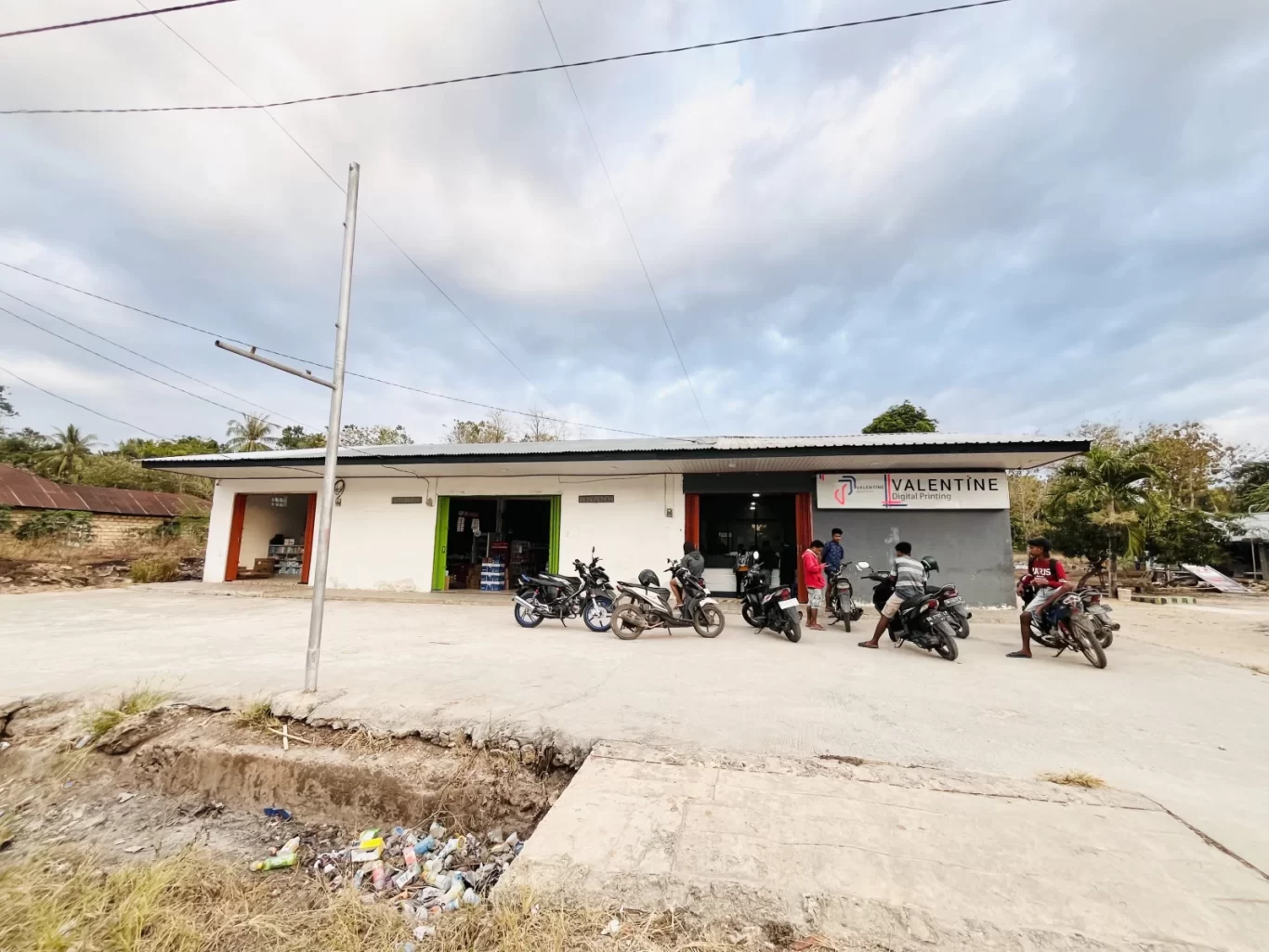 Valentine Digital Printing shop in Tambolaka, Sumba, with a white and grey facade, a wide concrete parking area, several motorbikes, and people.