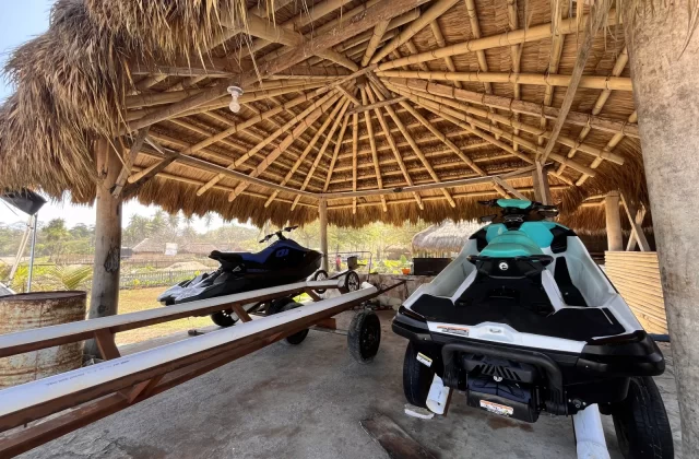 Two jet skis, one teal and white, the other dark blue, are parked under a large, round, thatched-roof hut with bamboo beams near a sunny beach.