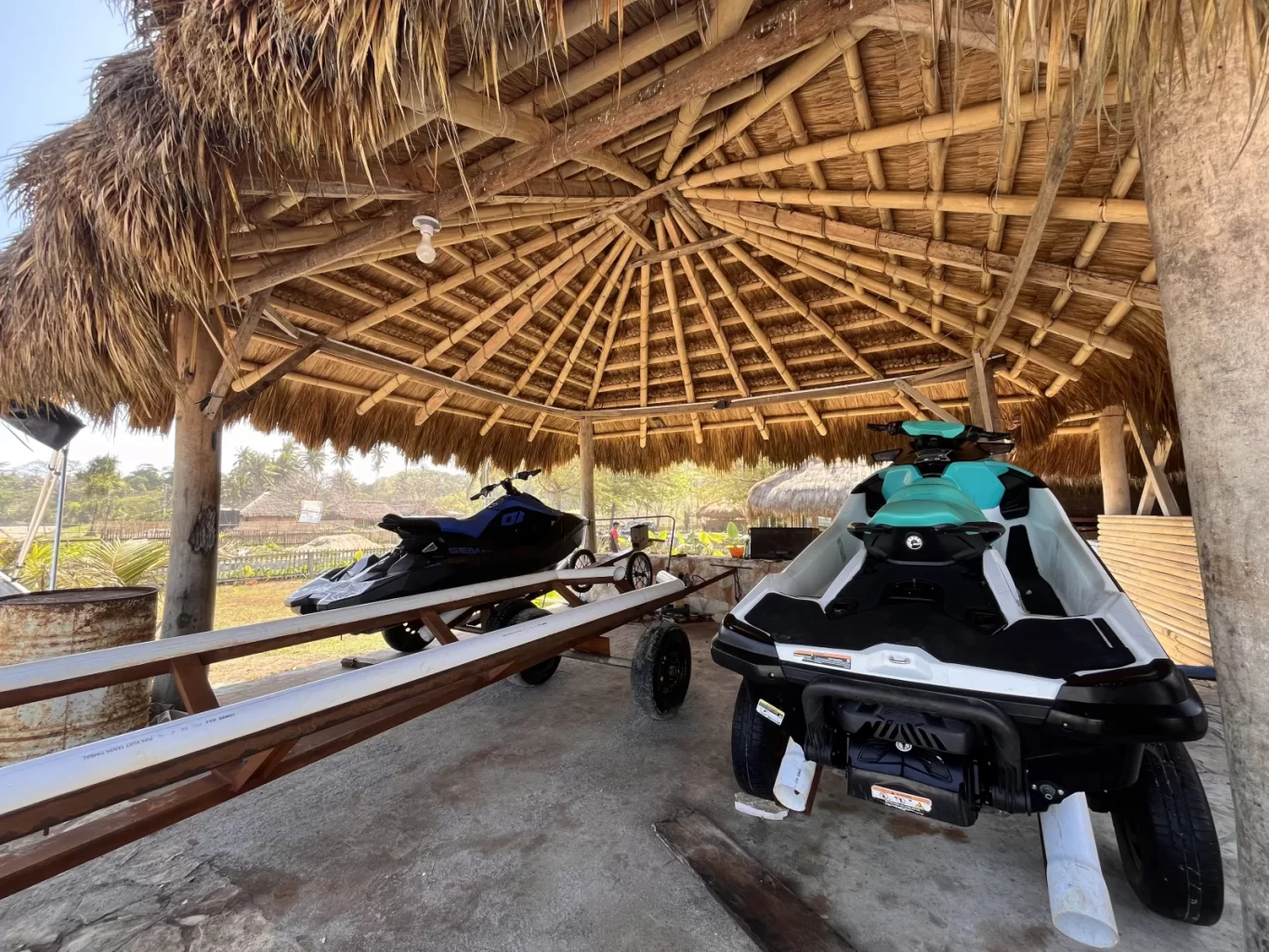 Two jet skis, one teal and white, the other dark blue, are parked under a large, round, thatched-roof hut with bamboo beams near a sunny beach.