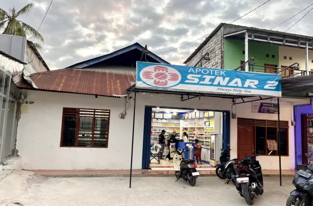 The white exterior of Sinar 2 Pharmacy in Tambolaka, Sumba, features a rusty brown roof and a blue sign, with motorbikes parked outside under a cloudy sky.