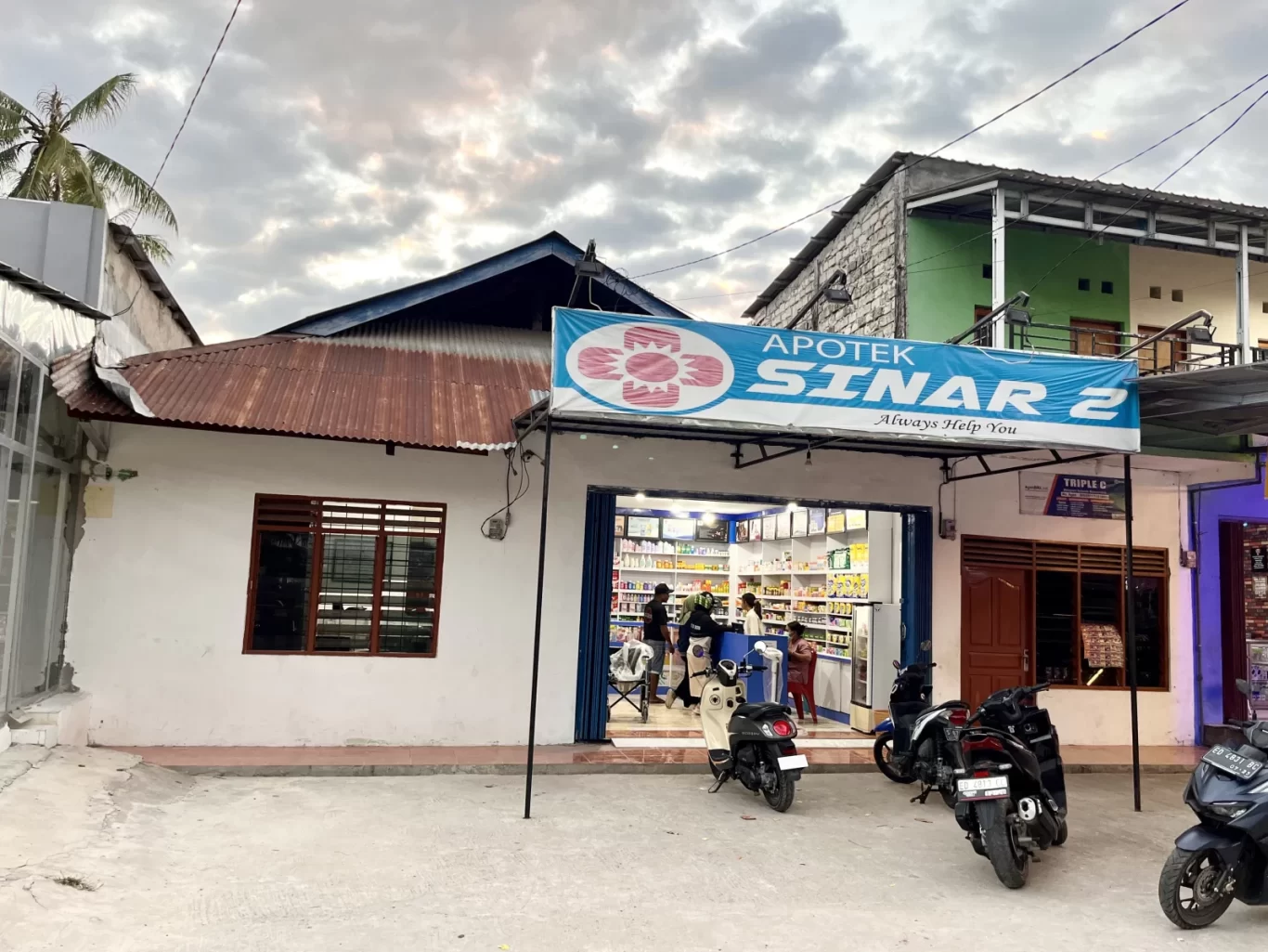 The white exterior of Sinar 2 Pharmacy in Tambolaka, Sumba, features a rusty brown roof and a blue sign, with motorbikes parked outside under a cloudy sky.