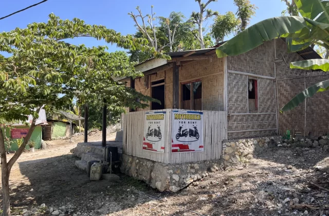 A simple, light brown bamboo-walled building with a "MOTORBIKE FOR RENT" sign, surrounded by green trees and dirt ground under a clear blue sky.