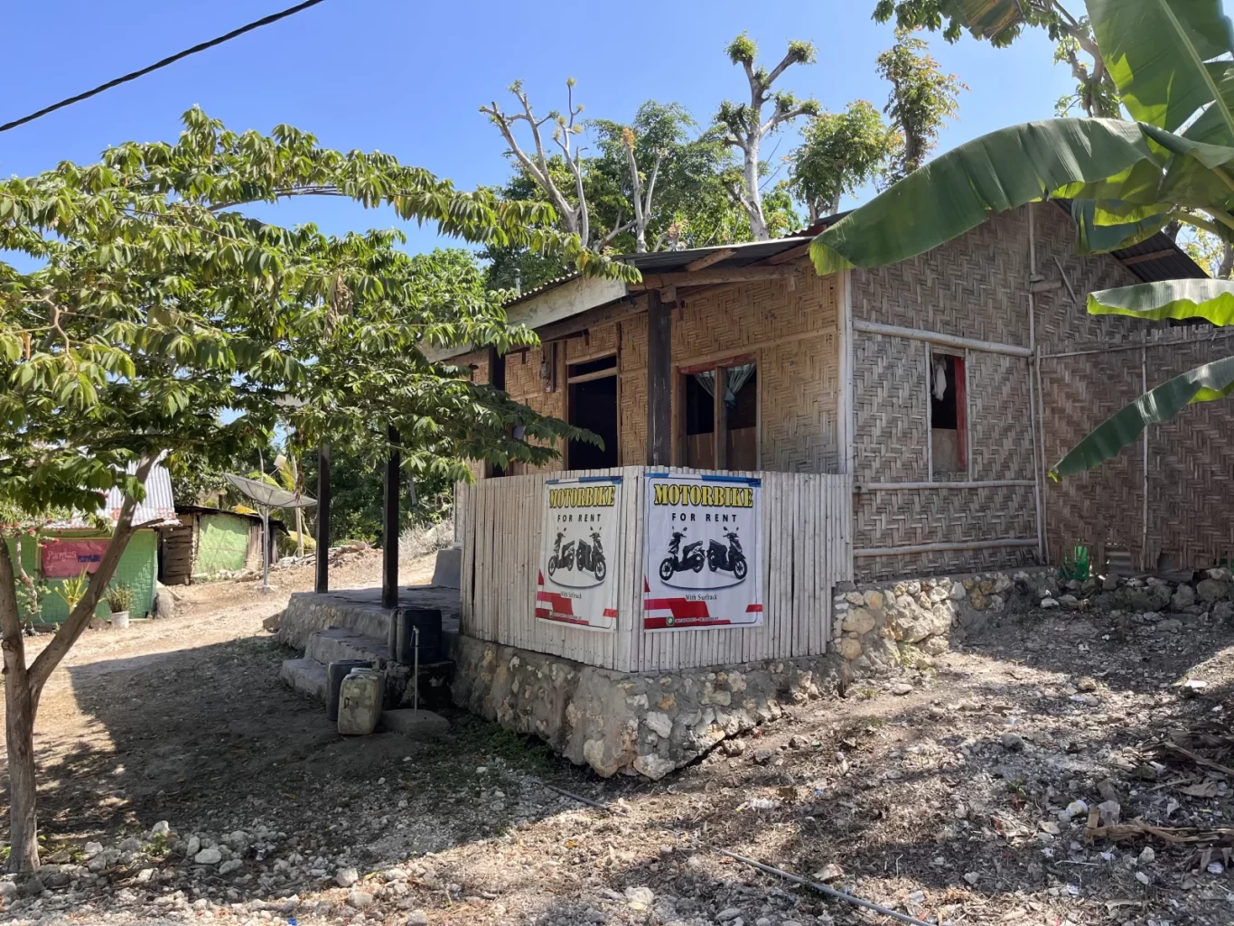 A simple, light brown bamboo-walled building with a "MOTORBIKE FOR RENT" sign, surrounded by green trees and dirt ground under a clear blue sky.
