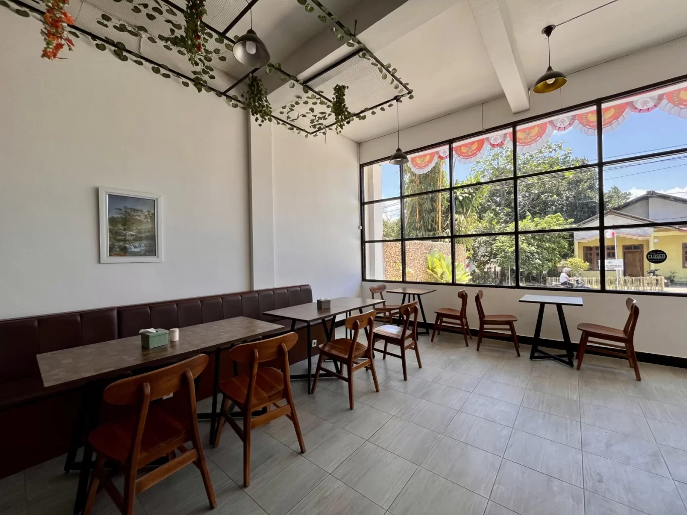 A bright, spacious restaurant interior features white walls, brown banquettes, wooden tables and chairs, and a large window overlooking lush trees.