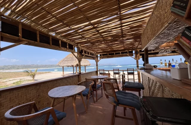 A bamboo and thatched roof restaurant with wooden tables and a bar overlooks Kerewe Beach and the blue ocean under a clear sky.