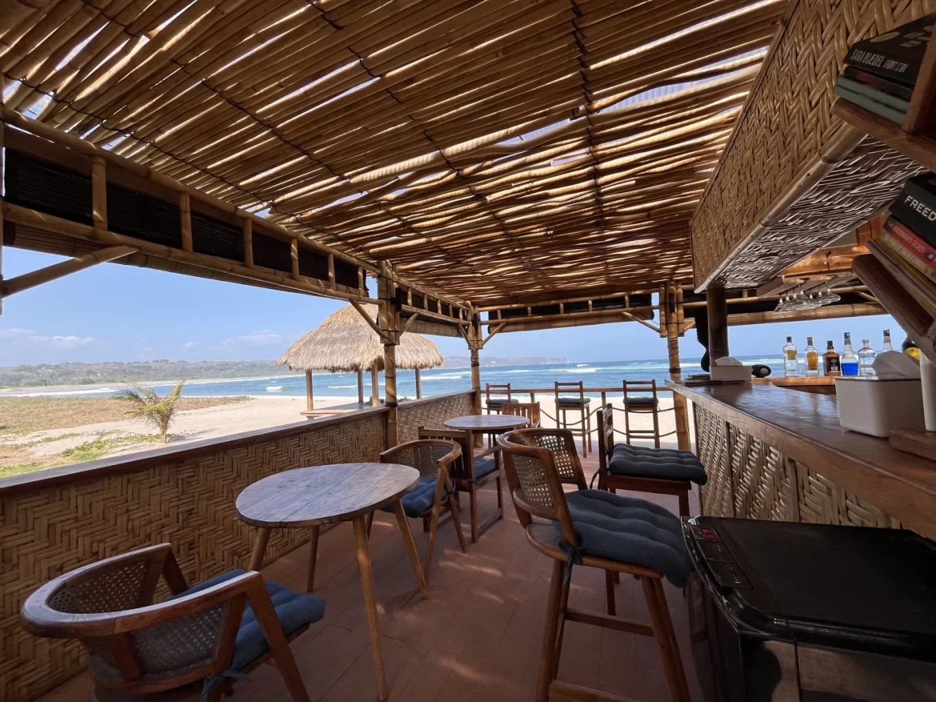 A bamboo and thatched roof restaurant with wooden tables and a bar overlooks Kerewe Beach and the blue ocean under a clear sky.