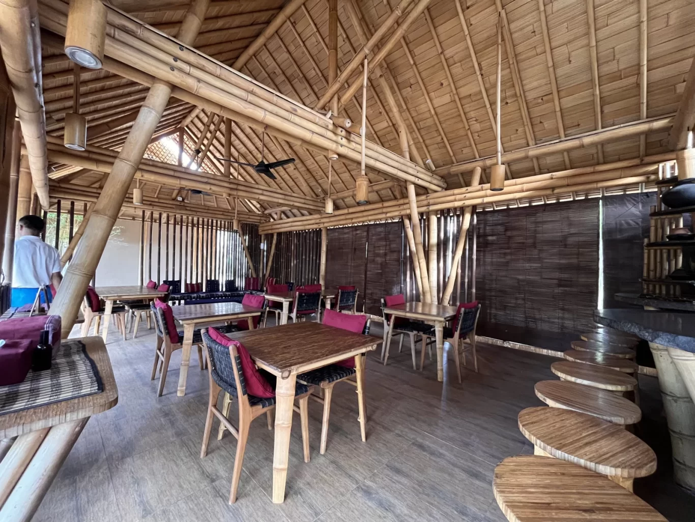 The interior of Maringi Restaurant features a high bamboo-beamed ceiling, wooden tables with red-cushioned chairs, and a bar with oval stools, with a person standing on the left.