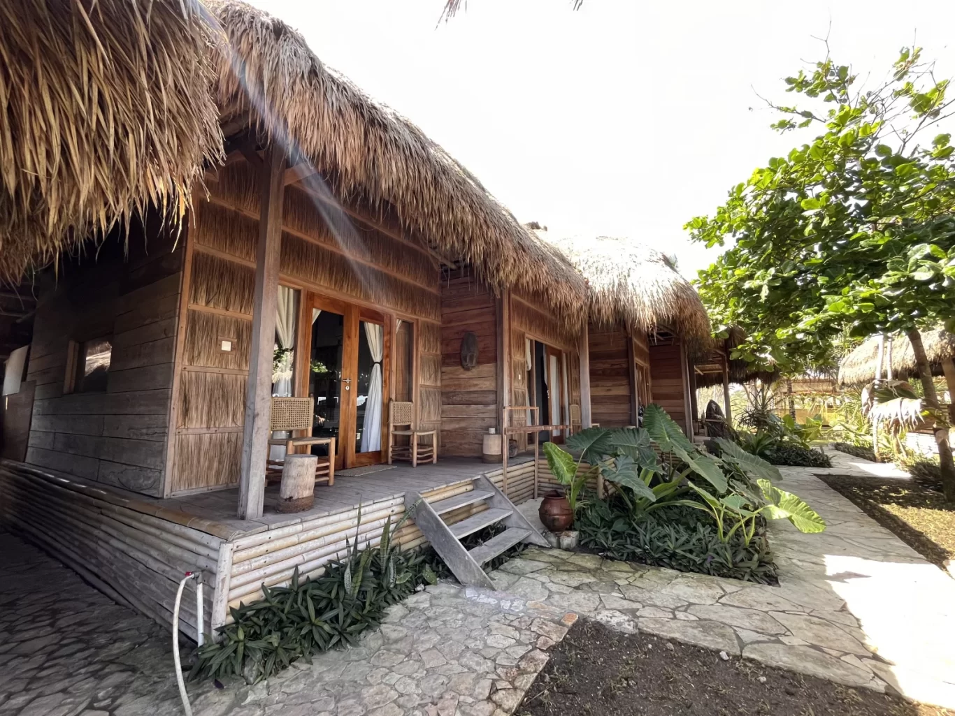 Traditional wooden bungalows with golden thatched roofs and private verandahs line a stone path, surrounded by lush green plants under a bright sky.