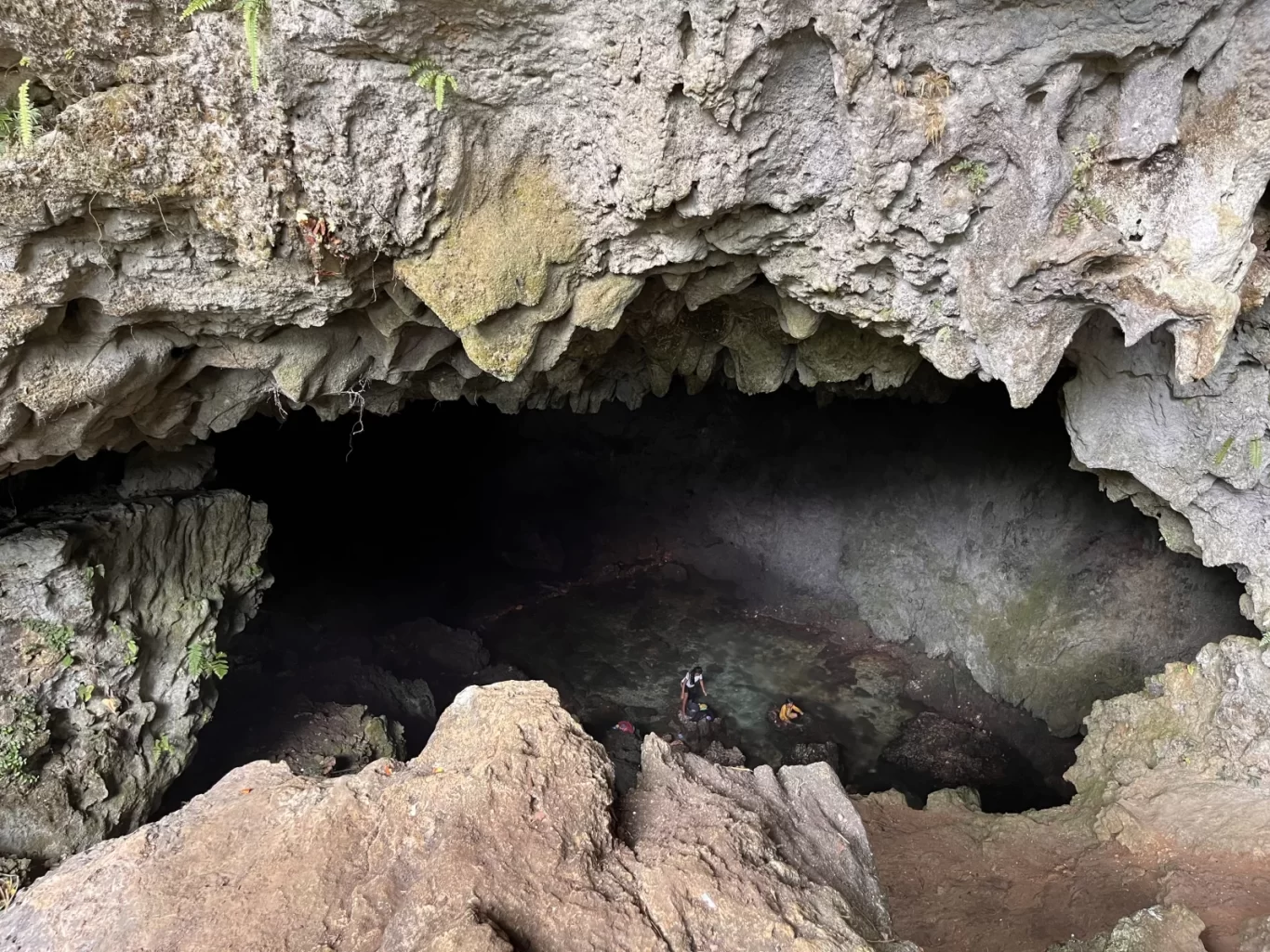The entrance of Gua Weli'a cave shows rugged grey rock formations, a clear spring, and two people exploring the dark interior.