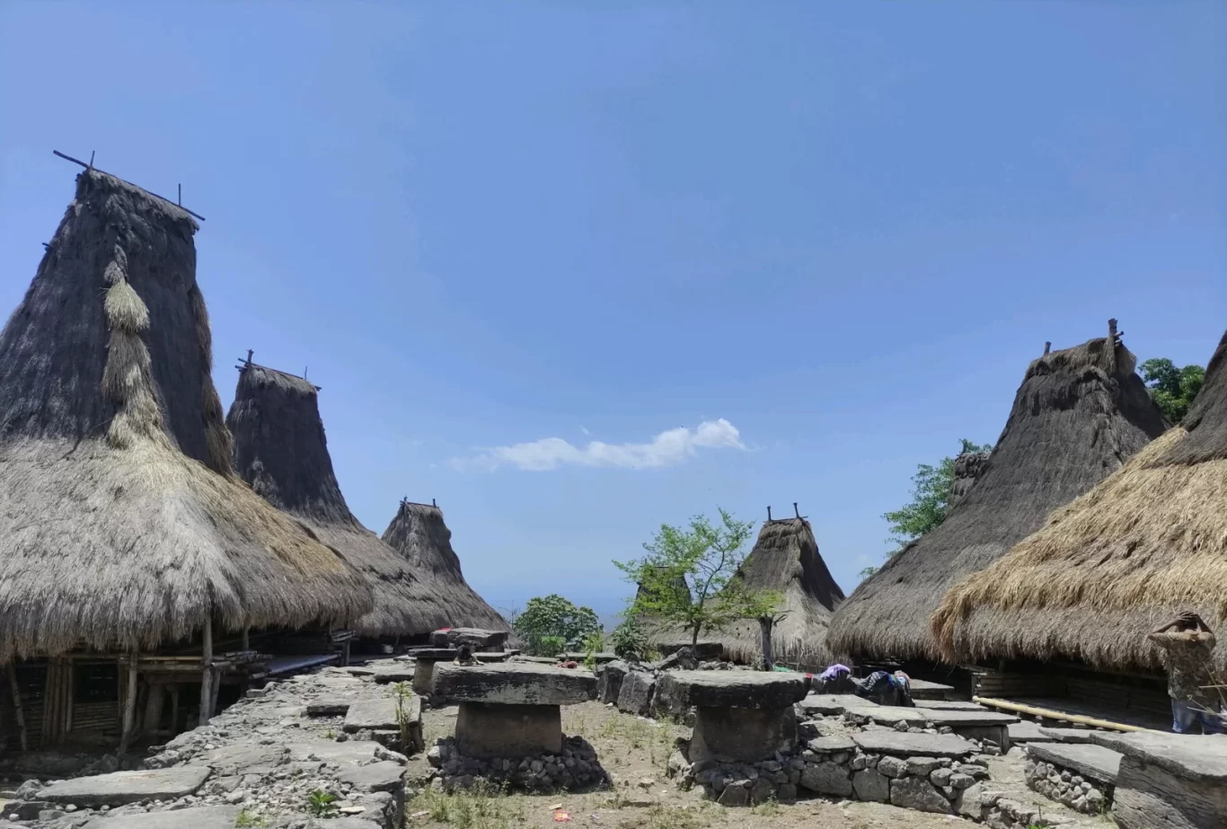 Traditional houses with tall, conical thatched roofs and ancient stone structures stand under a bright blue sky in Totok Kalada Village, Sumba.