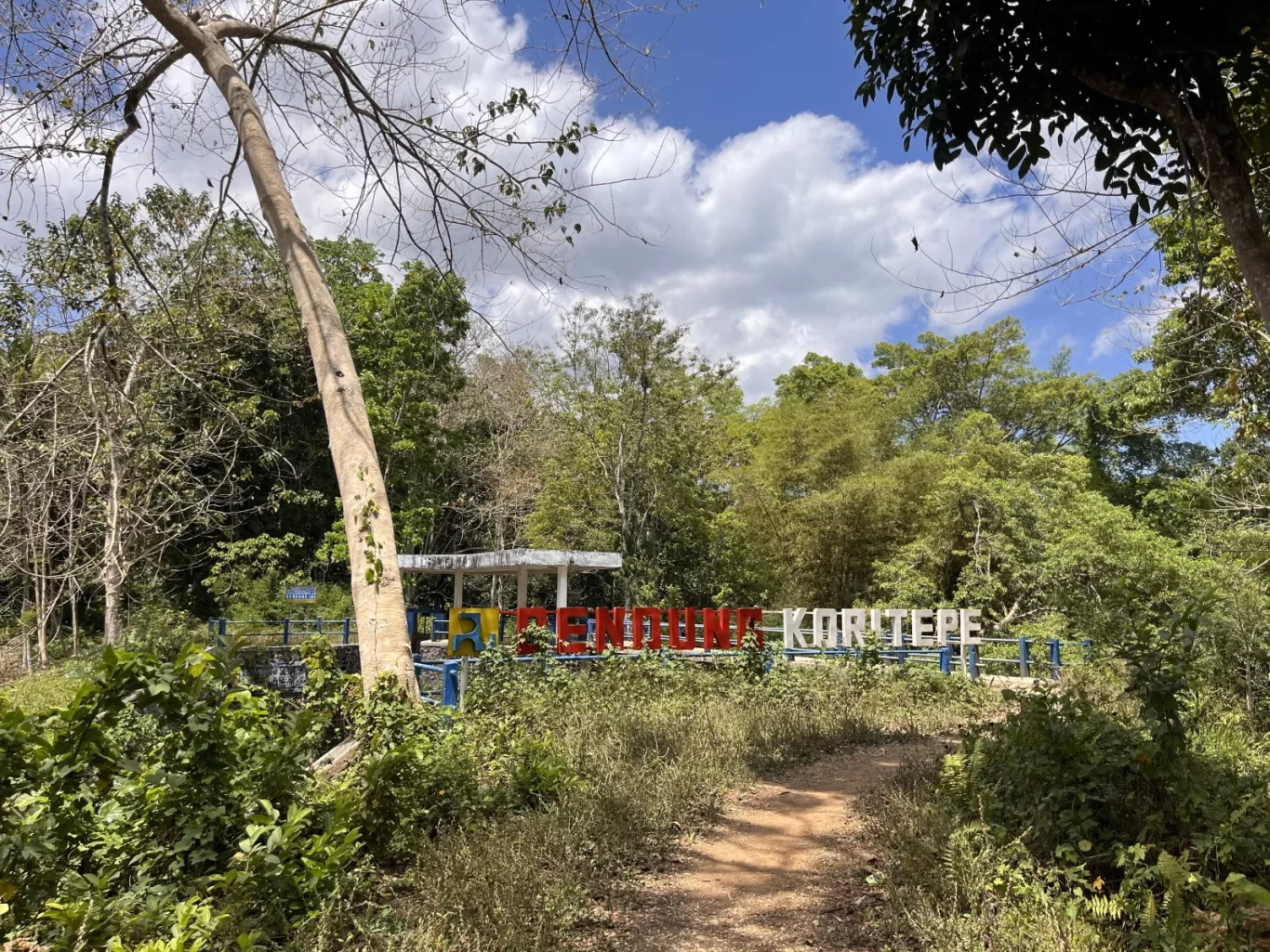 A dirt path winds through dense green foliage towards the Koritepe Dam, marked by a red and white sign on blue railings under a bright blue sky.