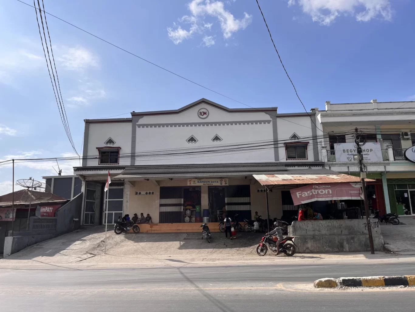 A white and gray two-story Bangun Jaya Mechanic shop stands under a blue sky in Tambolaka, Sumba, with motorcycles parked and people sitting outside.