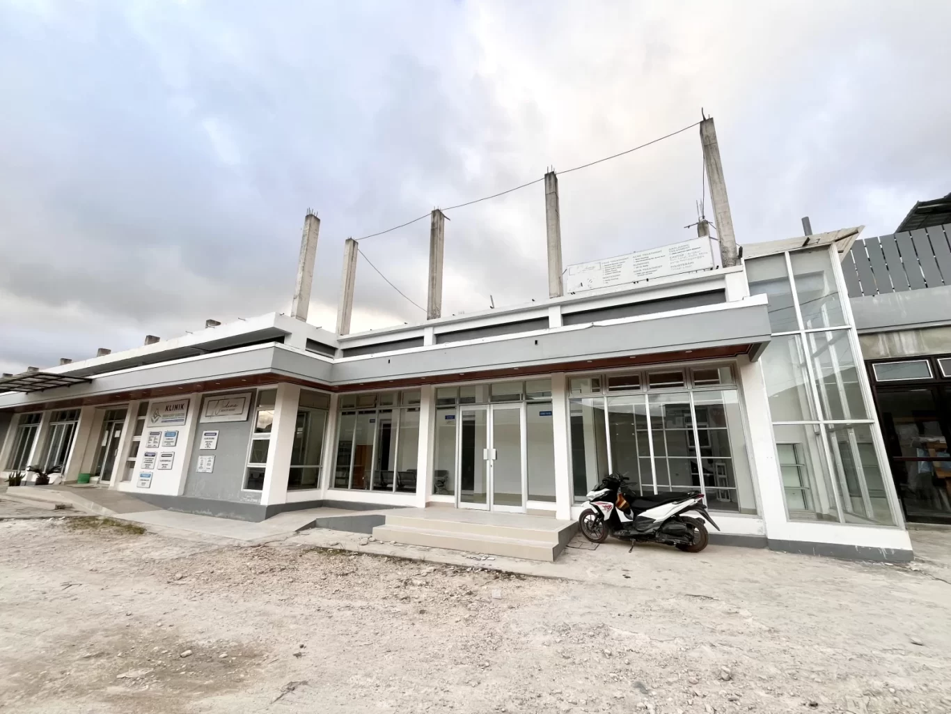 A modern grey and white building with large glass windows and doors stands under an overcast sky, with a white and black scooter parked in front.