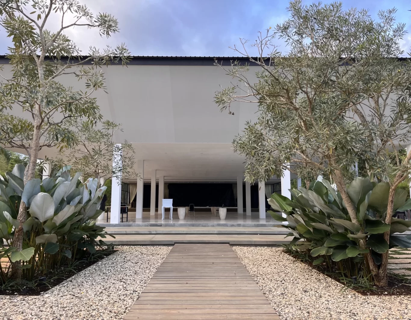 A modern white villa entrance features a wooden walkway flanked by gravel and lush tropical plants, leading to a columned porch under a partly cloudy sky.