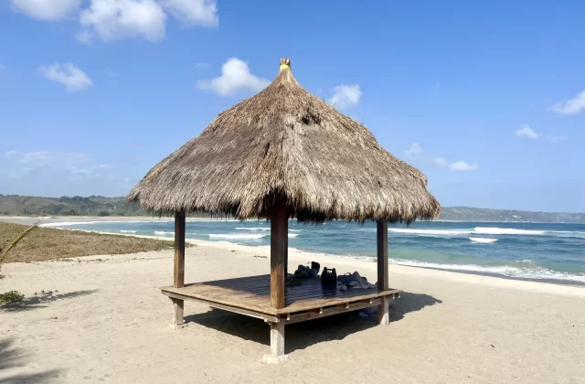 A traditional thatched-roof gazebo with a wooden platform stands on the sandy Kerewe Beach, overlooking the blue ocean under a clear sky.