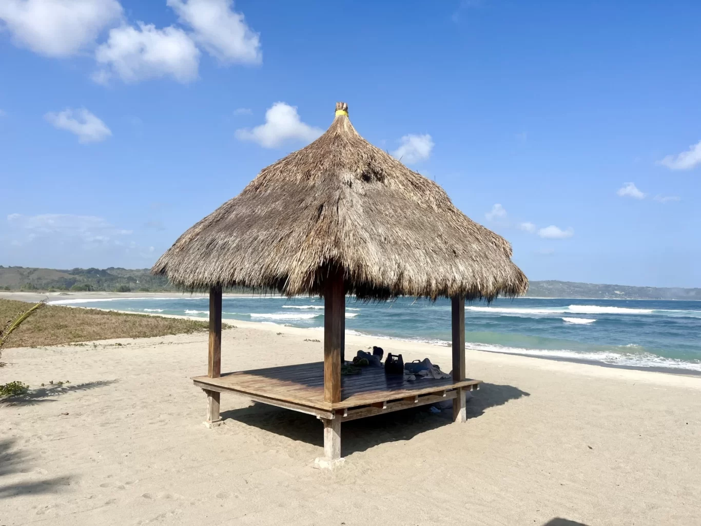 A traditional thatched-roof gazebo with a wooden platform stands on the sandy Kerewe Beach, overlooking the blue ocean under a clear sky.