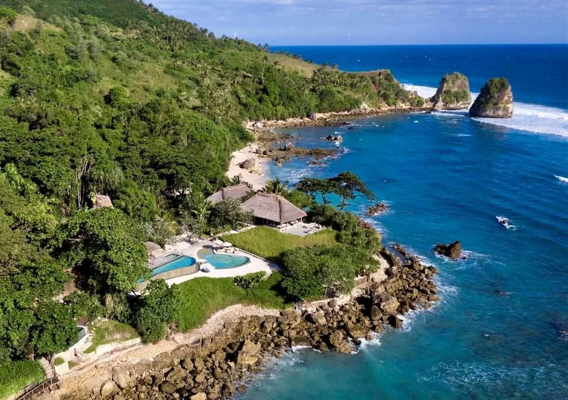 Aerial view of Villa Haweri, a complex with thatched roofs and pools, on a lush green, rocky coastline with deep blue ocean and sea stacks.