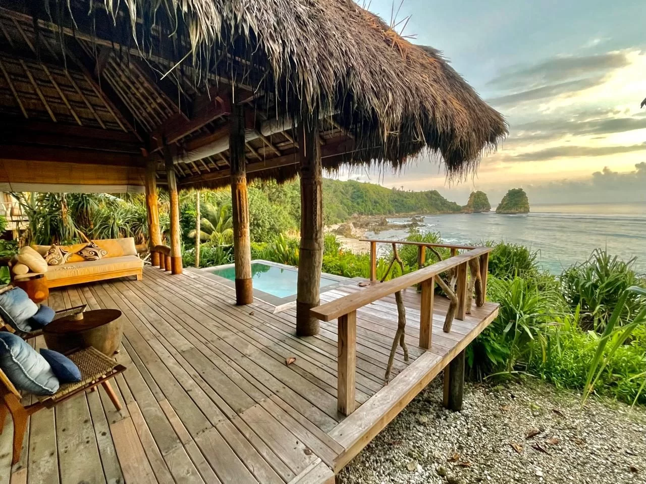 A thatched-roof villa patio with a wooden deck, seating, and a small plunge pool overlooks a tropical coastline with ocean, two rock formations, and a warm, cloudy sky.