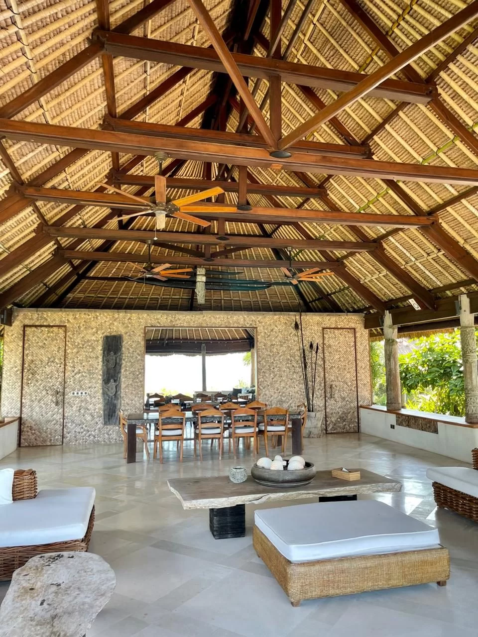 Open-air villa interior with a high thatched roof, dark wooden beams, and ceiling fans, featuring a dining area and a living space.