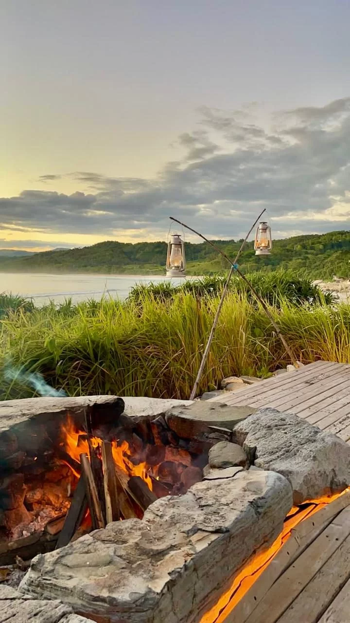 A lit bonfire on a deck, with two lanterns hanging over green coastal plants. An ocean and hills are visible under a golden, cloudy sky.