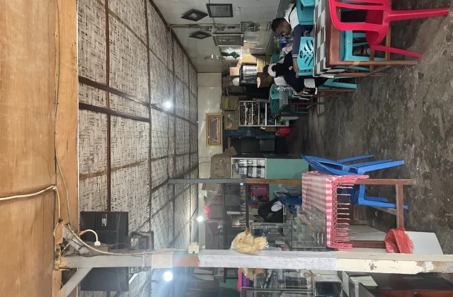The casual interior of Warung Arista in Waikabubak features woven bamboo walls, a concrete floor, and colorful plastic tables and chairs, with a few patrons seated.
