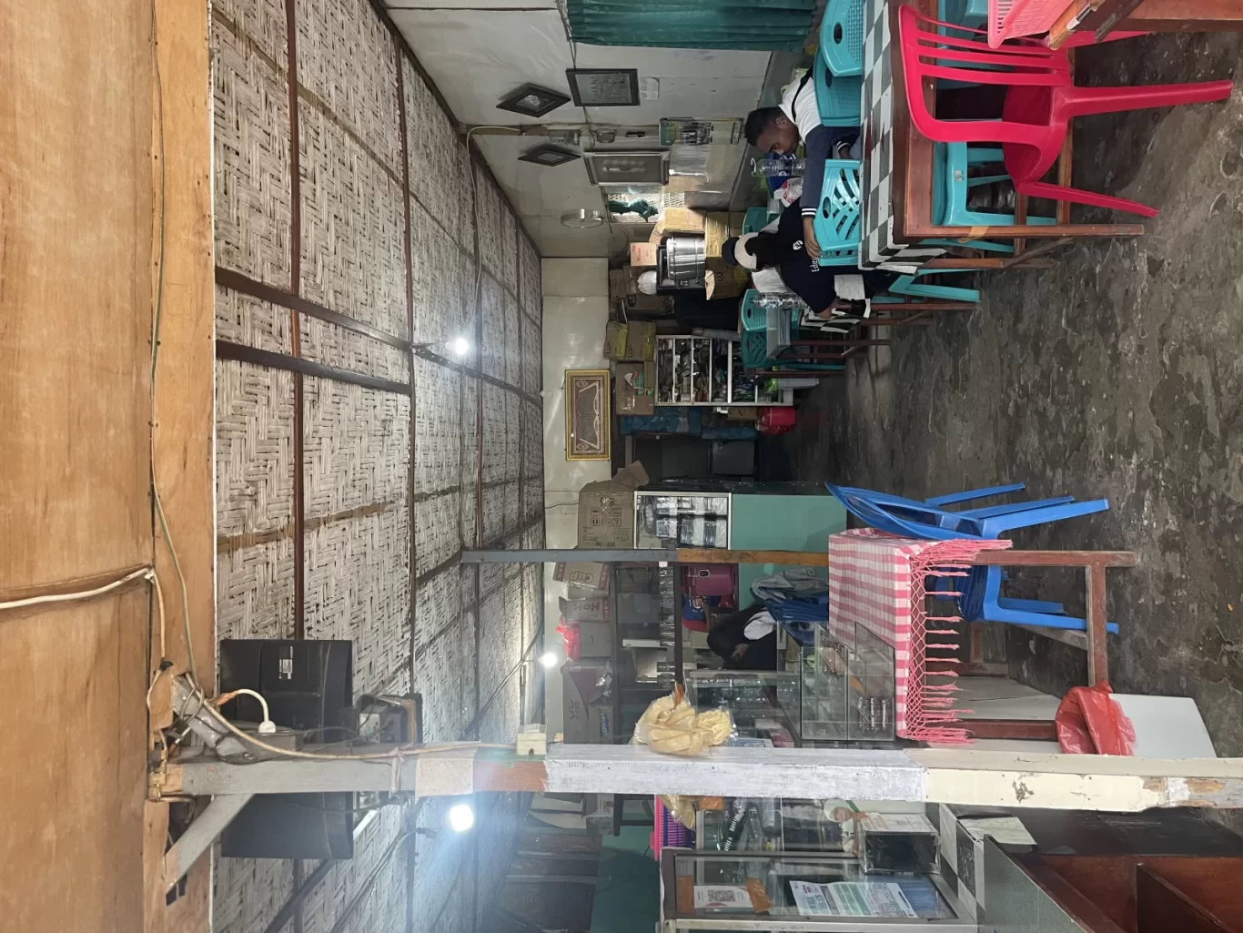 The casual interior of Warung Arista in Waikabubak features woven bamboo walls, a concrete floor, and colorful plastic tables and chairs, with a few patrons seated.