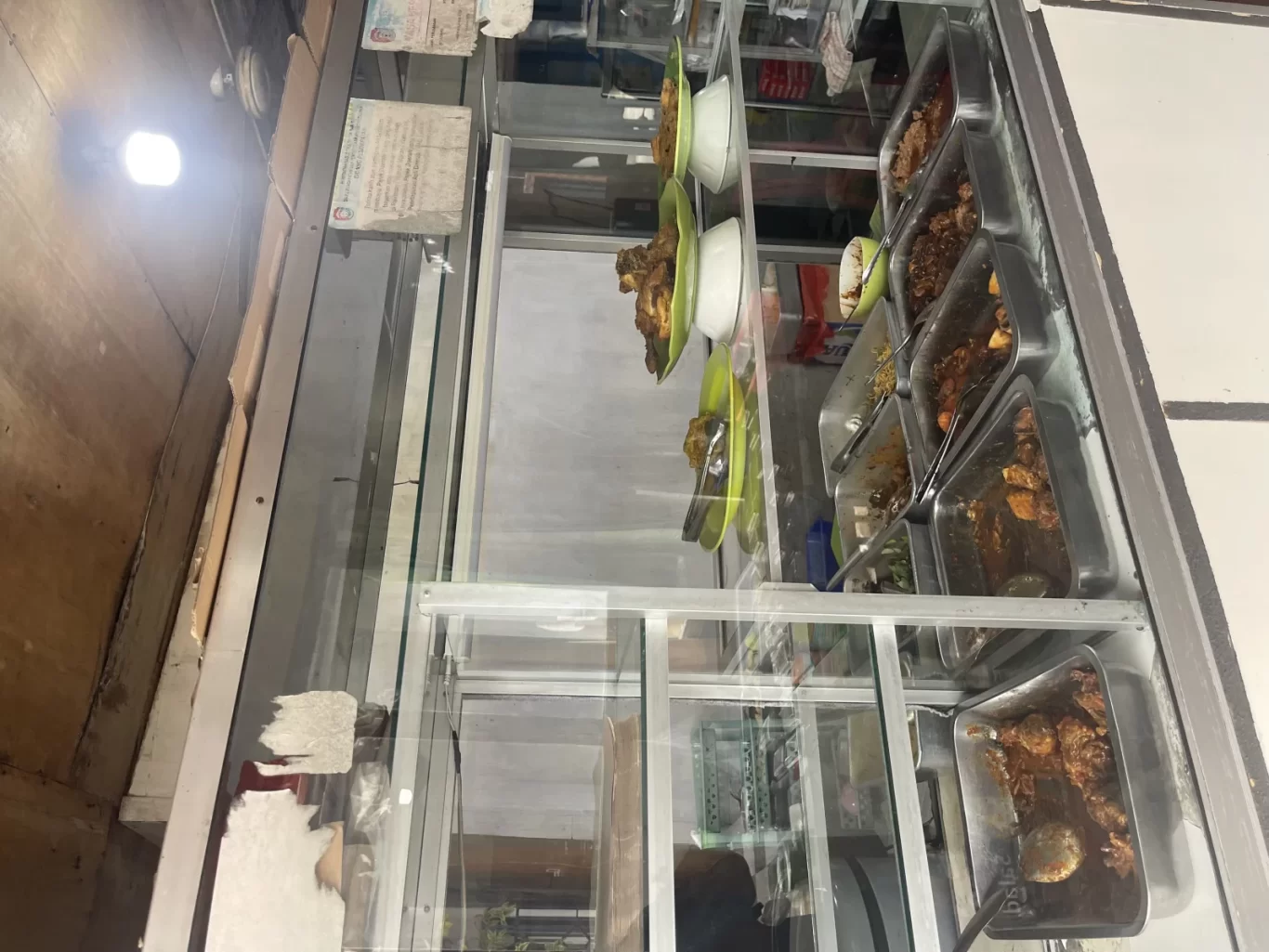 A glass display case at Warung Arista shows various Indonesian home-cooked dishes in metal trays and on green plates, brightly lit.