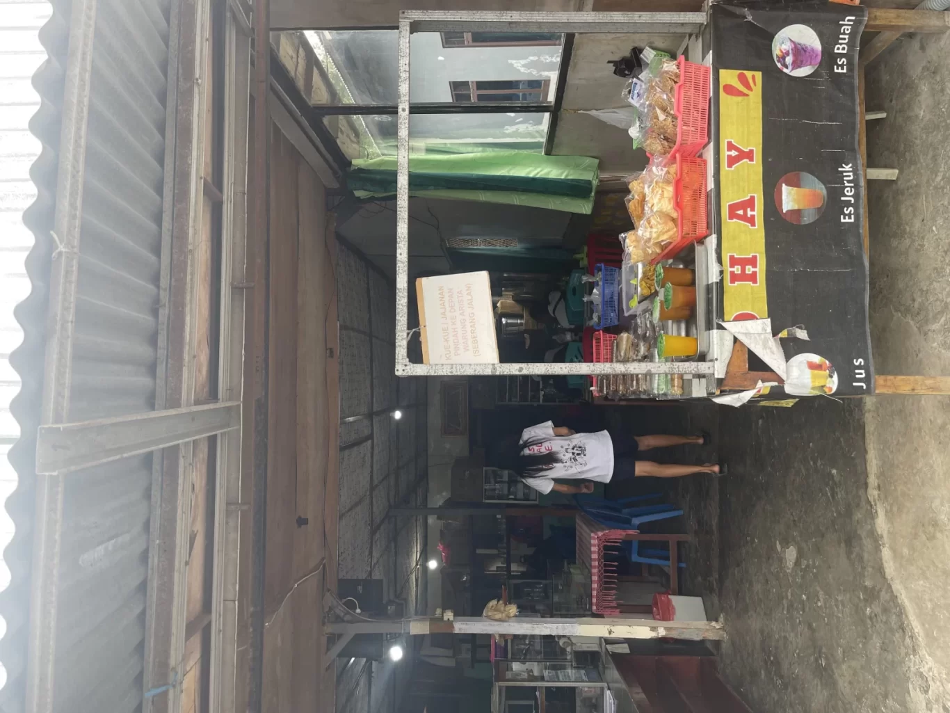 A woman stands inside a simple, open-fronted Warung Arista in Waikabubak, featuring a snack counter and a drink banner.