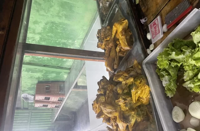 Golden-brown fried chicken pieces are displayed in a glass case next to a tray of fresh green lettuce and white cucumber slices at Warung Barokah.