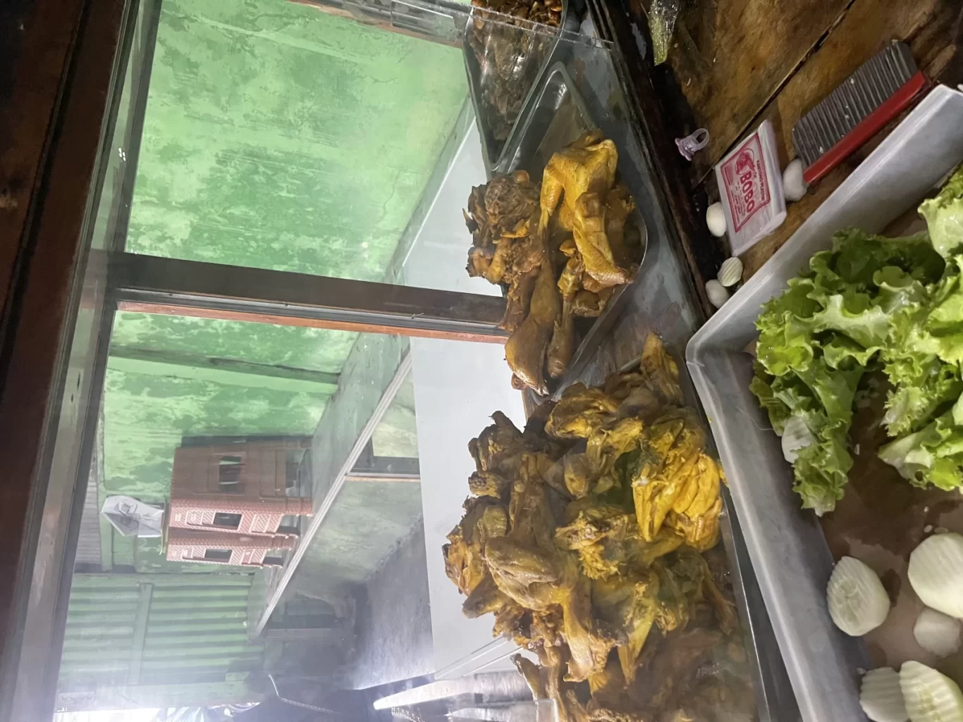 Golden-brown fried chicken pieces are displayed in a glass case next to a tray of fresh green lettuce and white cucumber slices at Warung Barokah.