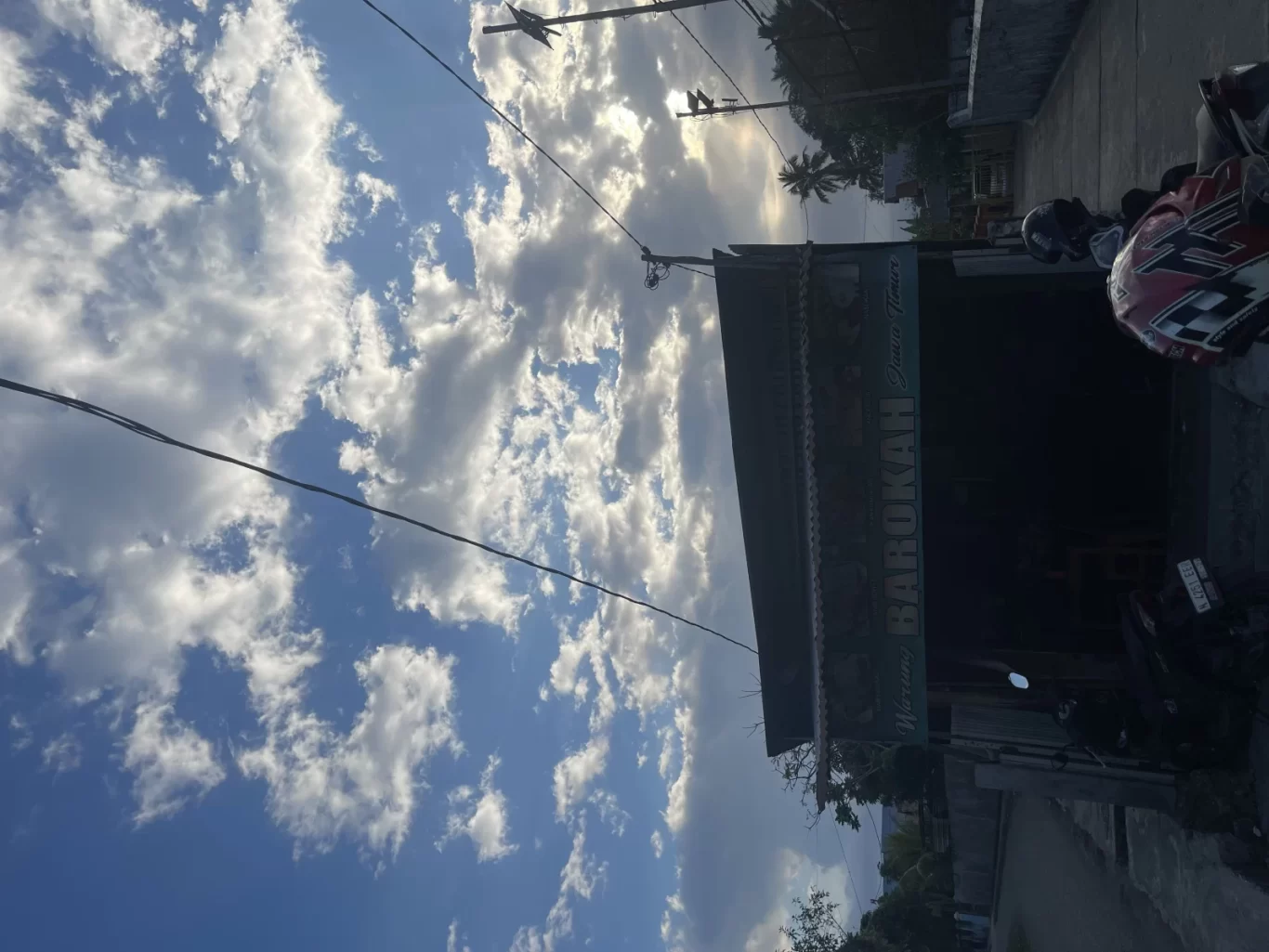 The exterior of Warung Barokah in Waikabubak, Sumba, is visible under a blue sky with white clouds and power lines. Motorcycles are parked in front of the dark building facade.