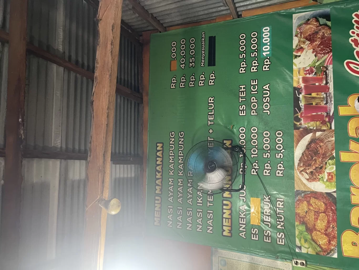 A green menu banner for Warung Barokah displays Indonesian food and drink prices, featuring images of fried fish, chicken, and colorful drinks. A small fan is attached, set against corrugated metal walls and wooden beams.