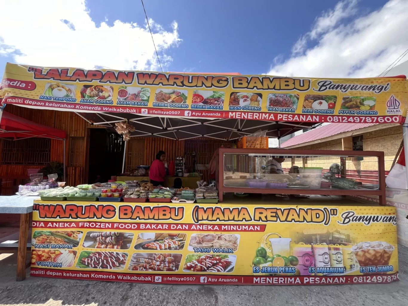 A vibrant yellow and red street food stall, Warung Bambu, with a vendor behind a counter displaying many Indonesian snacks under a blue sky.