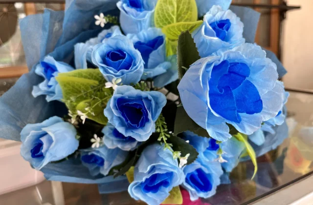 A vibrant bouquet of artificial blue roses with green leaves and small white filler flowers is wrapped in blue fabric, resting on a surface indoors.