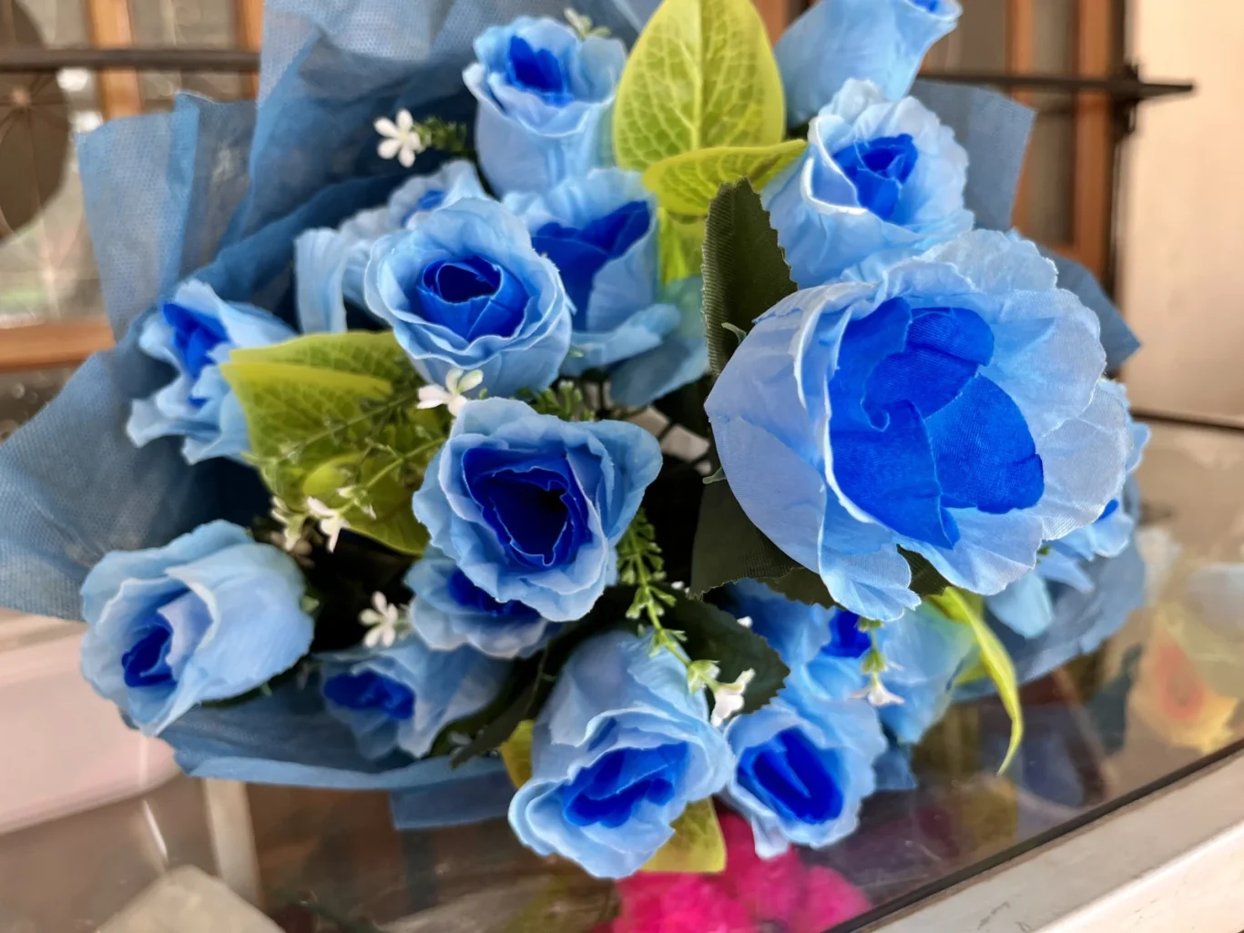 A vibrant bouquet of artificial blue roses with green leaves and small white filler flowers is wrapped in blue fabric, resting on a surface indoors.