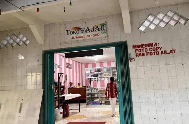 A person in a red and white shirt walks into the brightly lit "Toko Fajar" in Tambolaka, Sumba, with white tiled exterior walls and pink striped interior walls.