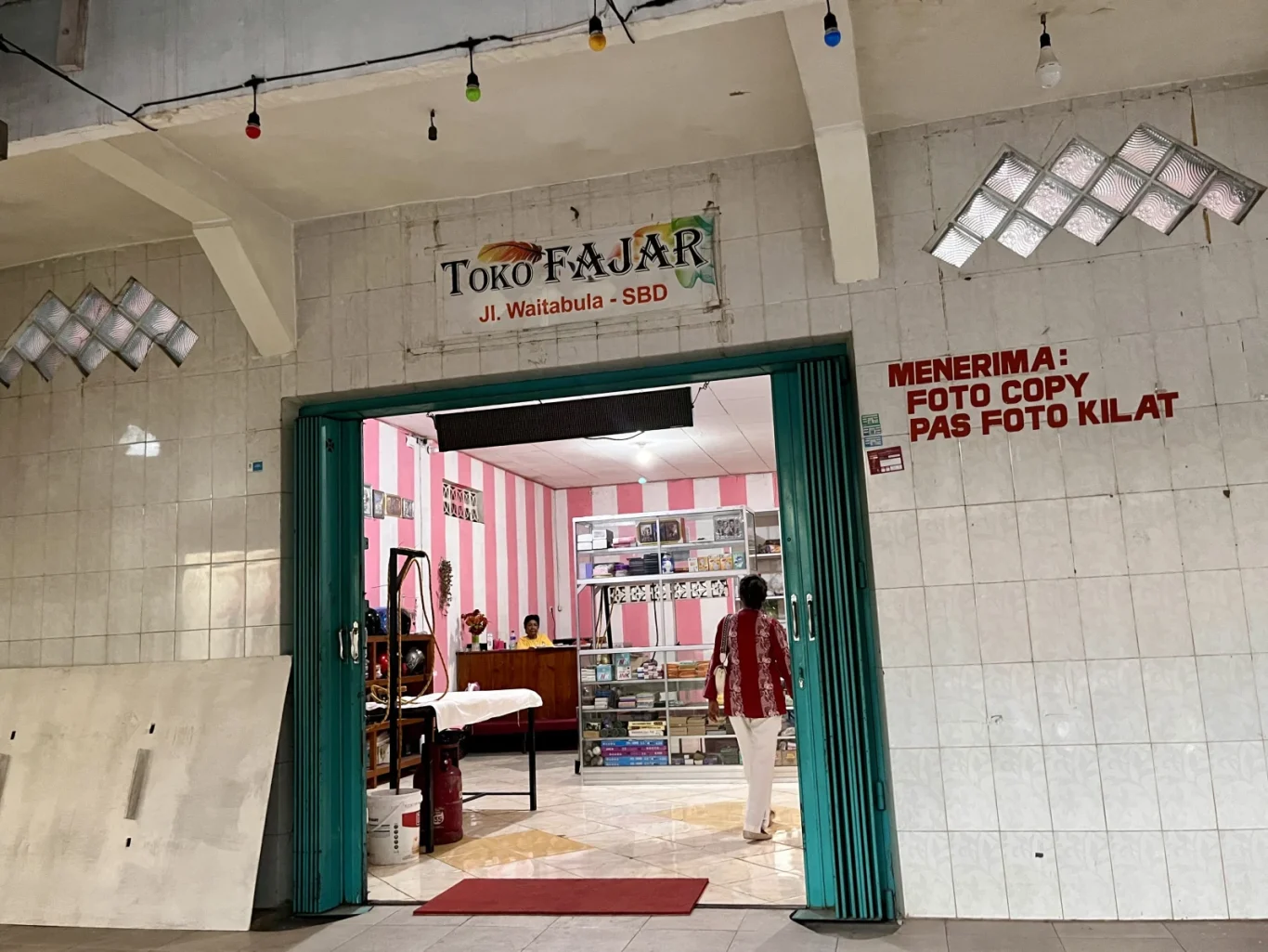 A person in a red and white shirt walks into the brightly lit "Toko Fajar" in Tambolaka, Sumba, with white tiled exterior walls and pink striped interior walls.
