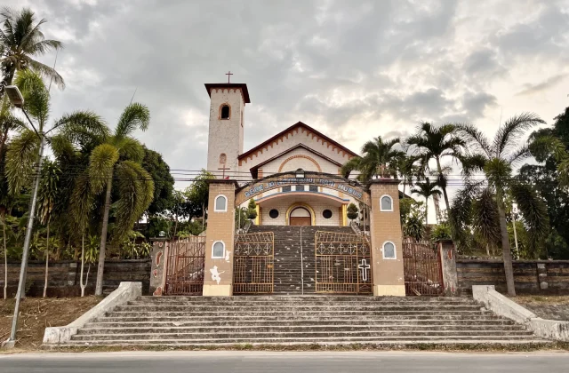 The Holy Spirit Cathedral Church in Tambolaka, Sumba, stands with a light-colored facade, bell tower, and ornate gates, flanked by palm trees under a cloudy sky.