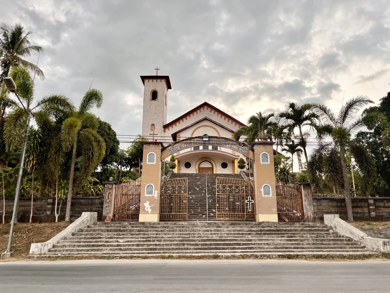 The Holy Spirit Cathedral Church in Tambolaka, Sumba, stands with a light-colored facade, bell tower, and ornate gates, flanked by palm trees under a cloudy sky.