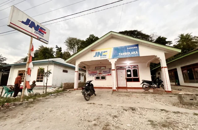 A white JNE Express building with a green roof stands on a dusty lot in Tambolaka, Sumba, under a cloudy sky. A large JNE sign and two men sitting on chairs are visible to the left, with a motorcycle parked in front.