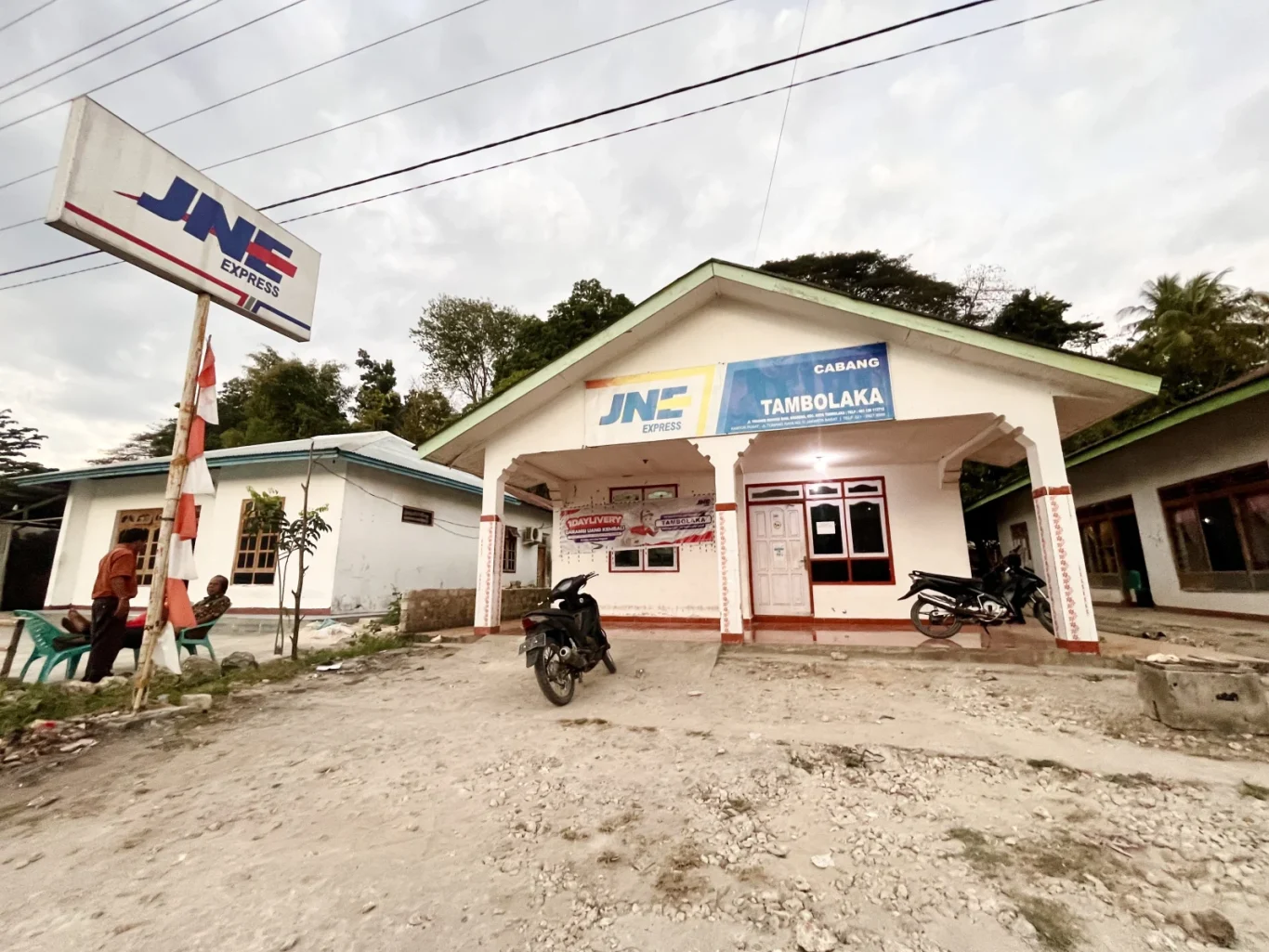 A white JNE Express building with a green roof stands on a dusty lot in Tambolaka, Sumba, under a cloudy sky. A large JNE sign and two men sitting on chairs are visible to the left, with a motorcycle parked in front.