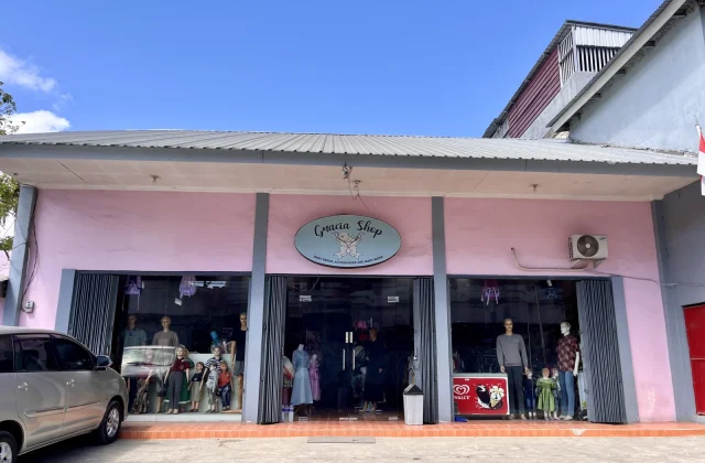The pink Gracia Shop building with a grey roof stands under a blue sky, displaying mannequins in its large glass windows, with a silver car parked on the left.