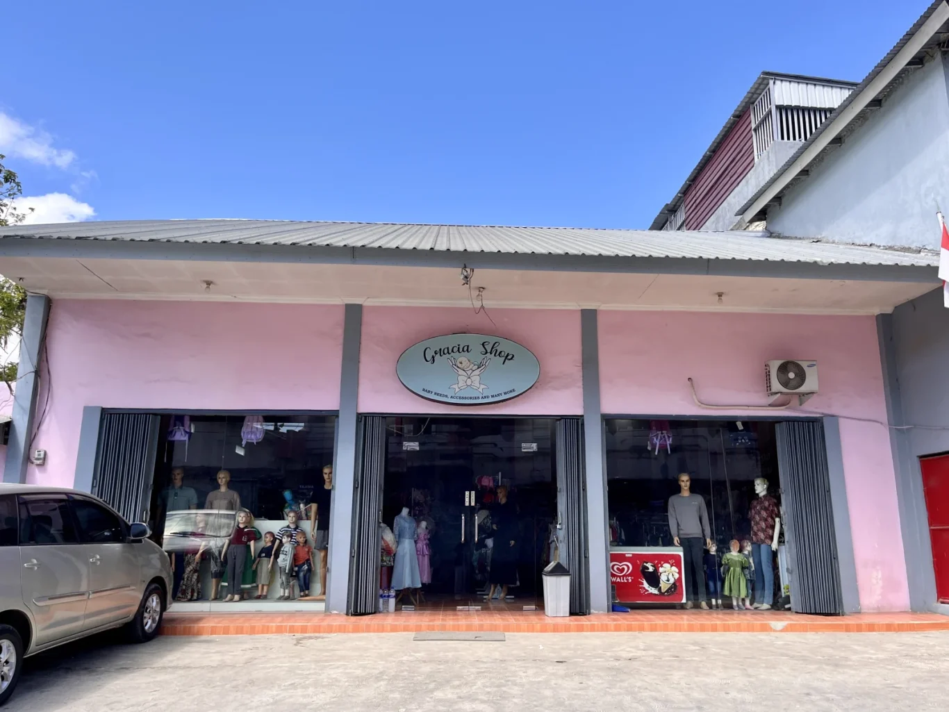 The pink Gracia Shop building with a grey roof stands under a blue sky, displaying mannequins in its large glass windows, with a silver car parked on the left.