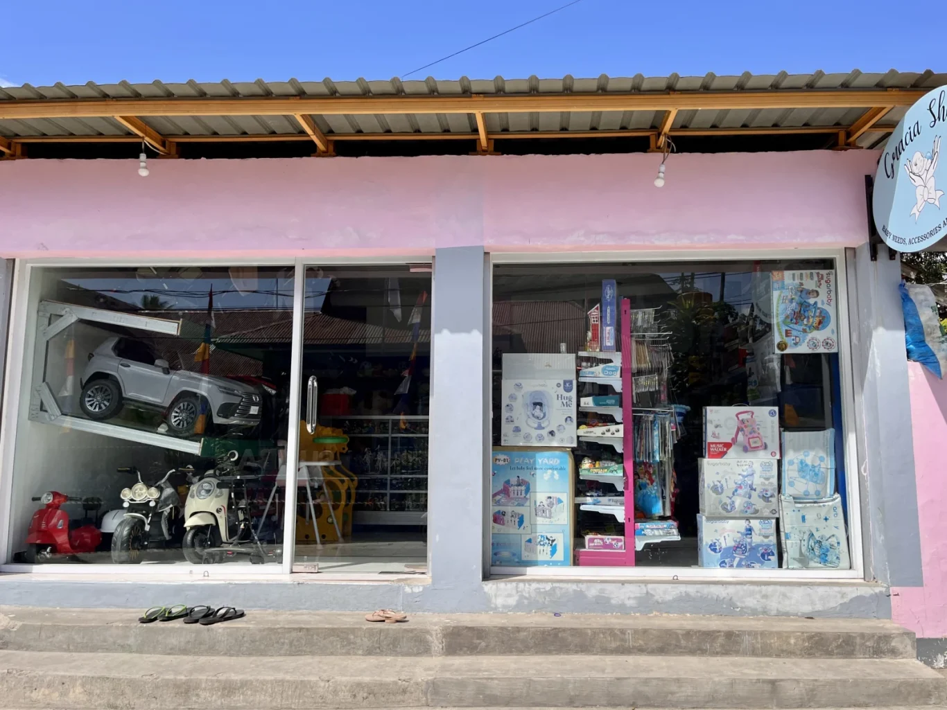 Grace Toy Shop in Waikabubak, with a pink facade, displays toy vehicles and boxed baby items in its large glass windows under a clear blue sky.