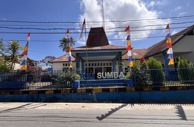 The white and blue RRI Sumba building with a traditional roof stands behind a blue fence adorned with Indonesian and colorful flags under a bright blue sky.